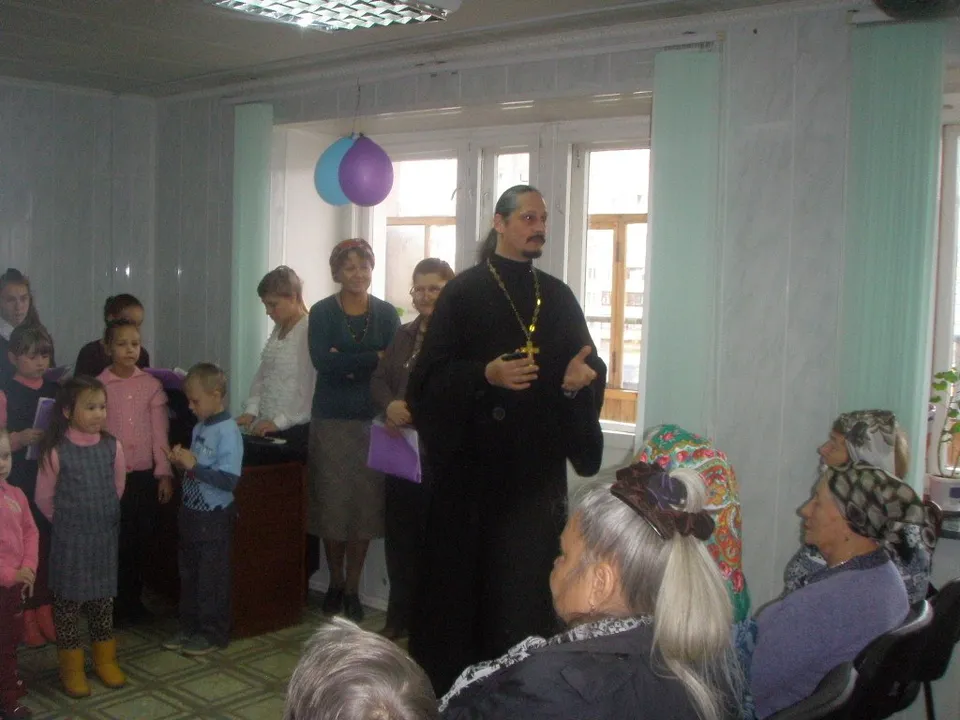 Священник беседует с детьми и пожилыми людьми в зале