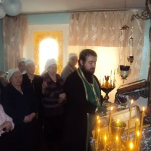 Священник в зелёном облачении совершает каждение у аналоя со свечами; рядом стоят пожилые прихожанки в платках, на заднем плане окно с занавесками молебной комнаты